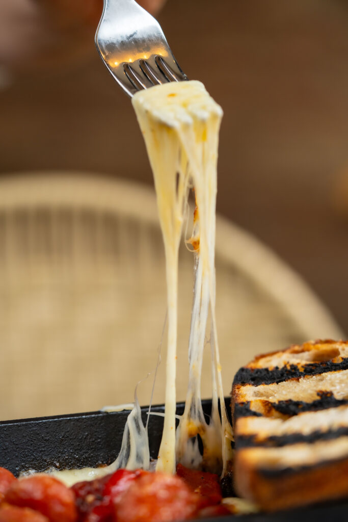 A dramatic shot of melted cheese stretching upward as it’s lifted from a sizzling cast iron skillet, paired with grilled bread and a tomato-based accompaniment below.
