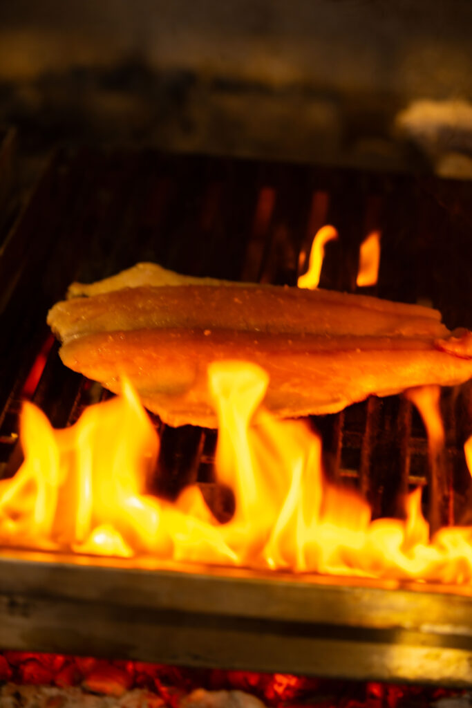 A fish fillet is being grilled over open flames on a wood-fired grill. The golden-brown skin and vivid flames evoke a sense of smoky flavor and traditional cooking over fire.